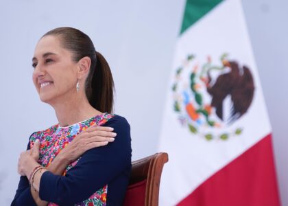 Claudia Sheinbaum sonríe sentada junto a la bandera de México durante el acto.